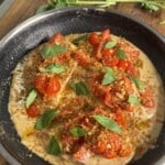 A skillet with creamy sauce topped with chopped cherry tomatoes, fresh basil leaves, and golden breadcrumbs sits on a wooden surface, with sprigs of fresh basil nearby.