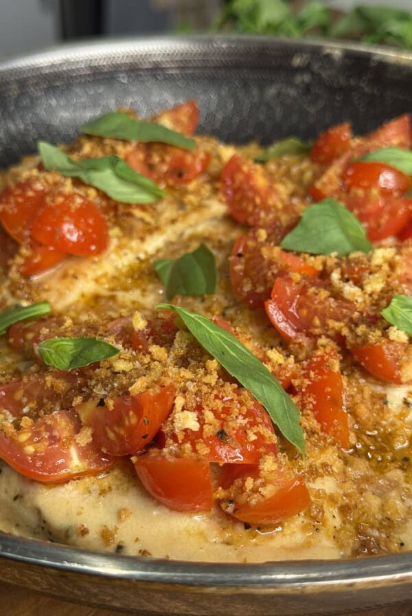 A close-up of a pan with cooked chicken breasts topped with chopped cherry tomatoes, crispy breadcrumbs, and fresh basil leaves. The dish appears juicy and garnished, ready to serve.