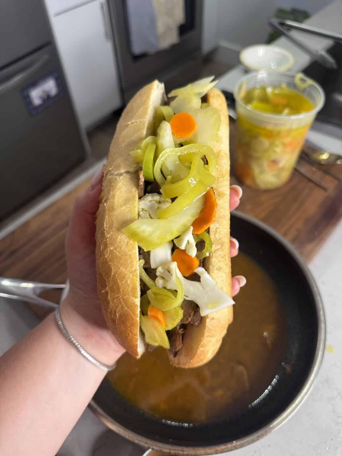 A hand holds an Italian beef sandwich in a crusty roll, topped with sliced peppers, carrots, and giardiniera, over a pan of broth. A container of more giardiniera sits on the counter in the background.