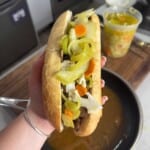A hand holds an Italian beef sandwich in a crusty roll, topped with sliced peppers, carrots, and giardiniera, over a pan of broth. A container of more giardiniera sits on the counter in the background.
