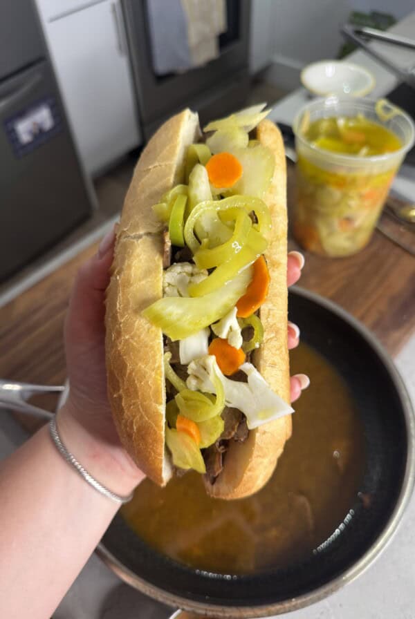 A hand holds an Italian beef sandwich in a crusty roll, topped with sliced peppers, carrots, and giardiniera, over a pan of broth. A container of more giardiniera sits on the counter in the background.