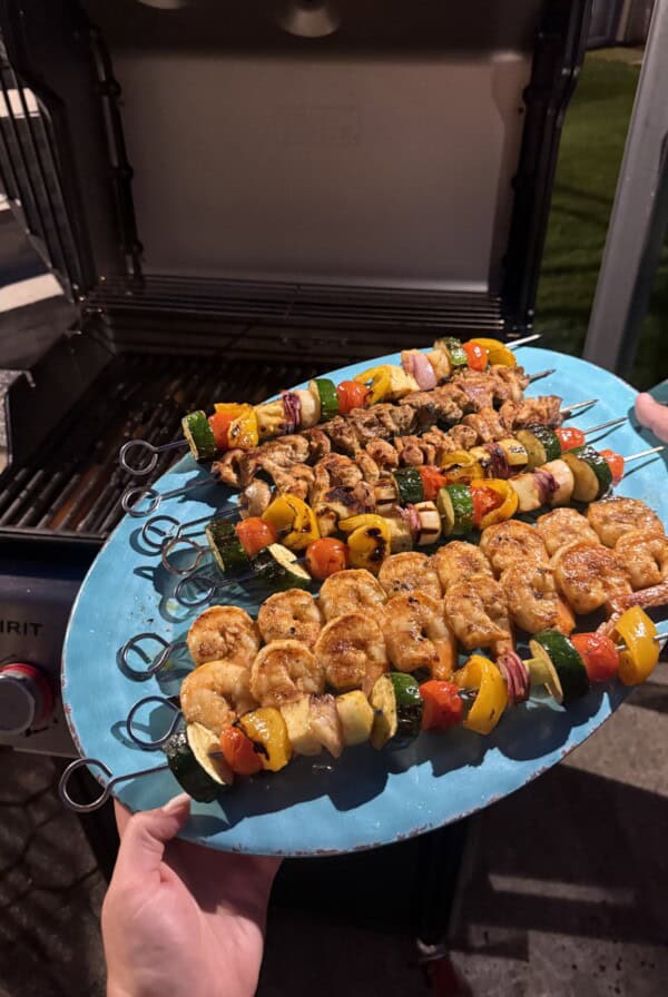 A person holds a blue platter with colorful skewers of shrimp, chicken, and vegetables, ready to be grilled on an open barbecue grill at night.