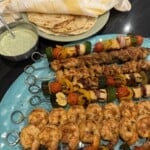 A platter of grilled kebabs with shrimp, chicken, vegetables, and paneer, served with flatbreads, a bowl of green sauce, and a cloth napkin on a plate in the background.