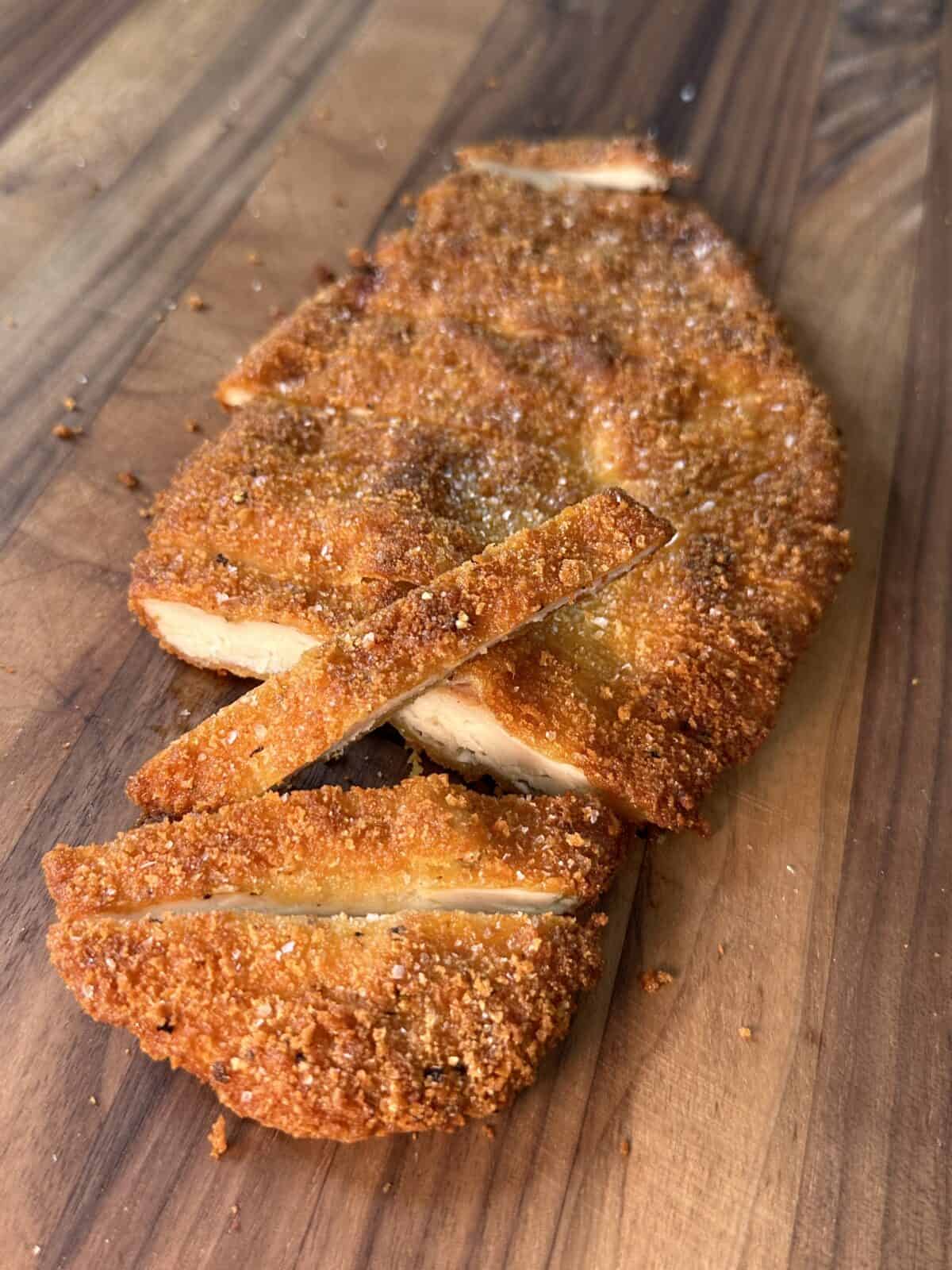 A breaded and fried cutlet, sliced into strips, sits on a wooden surface. The golden-brown crust appears crispy and the interior reveals juicy, cooked meat.