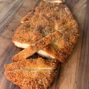 A breaded and fried cutlet, sliced into strips, sits on a wooden surface. The golden-brown crust appears crispy and the interior reveals juicy, cooked meat.