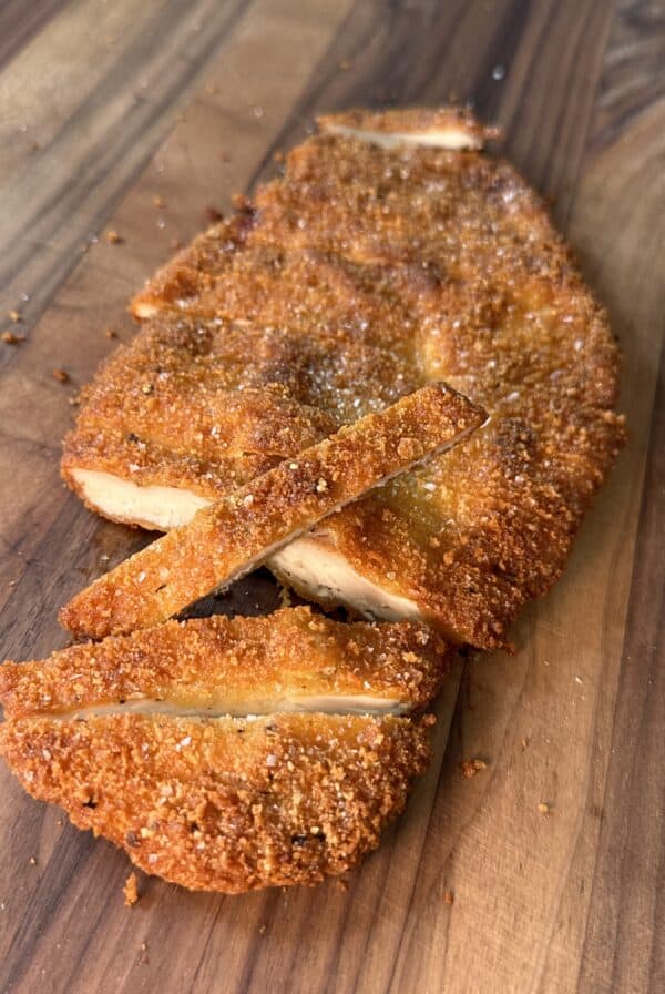 A breaded and fried cutlet, sliced into strips, sits on a wooden surface. The golden-brown crust appears crispy and the interior reveals juicy, cooked meat.
