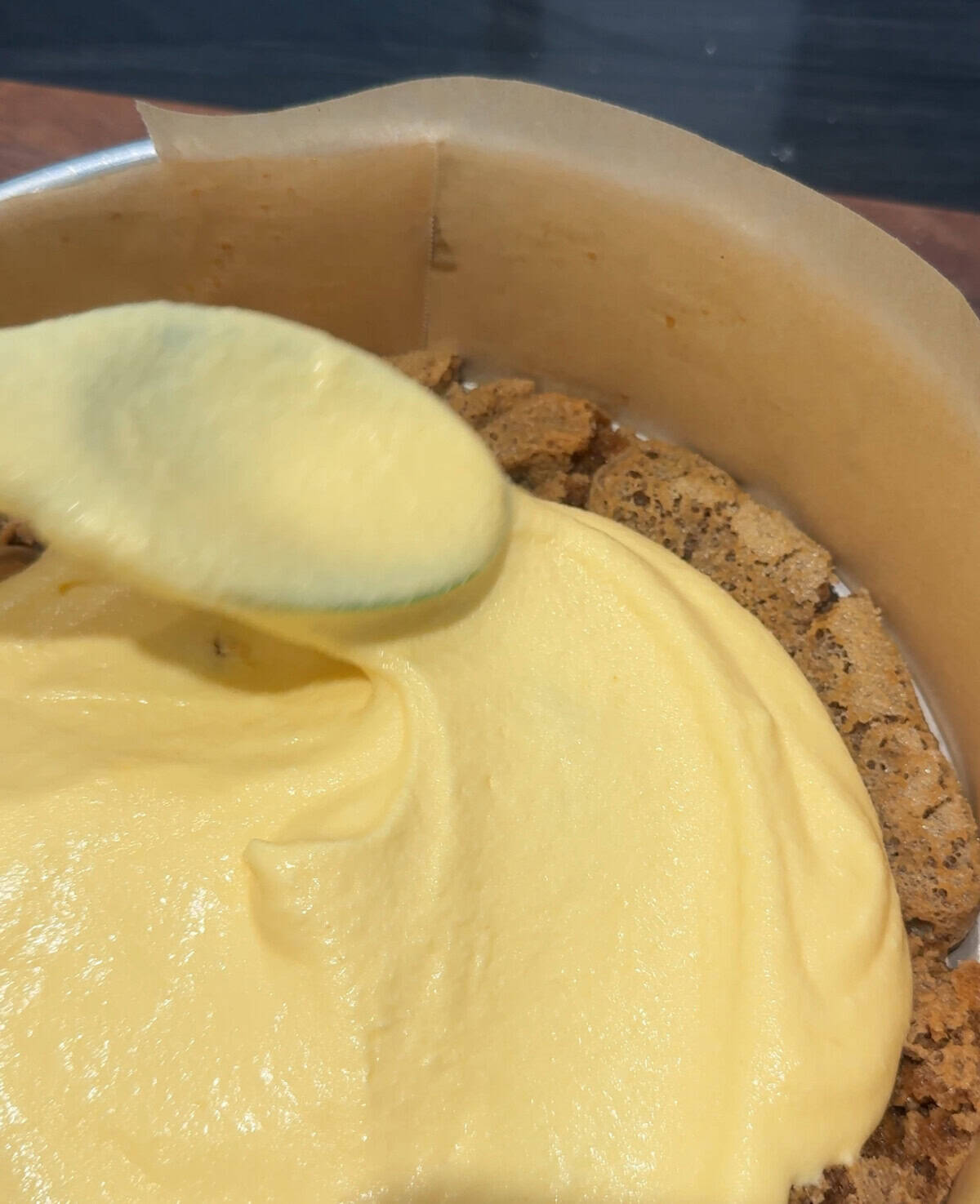 A spoon spreads creamy yellow filling over a crumbly cookie crust in a parchment-lined cake pan.