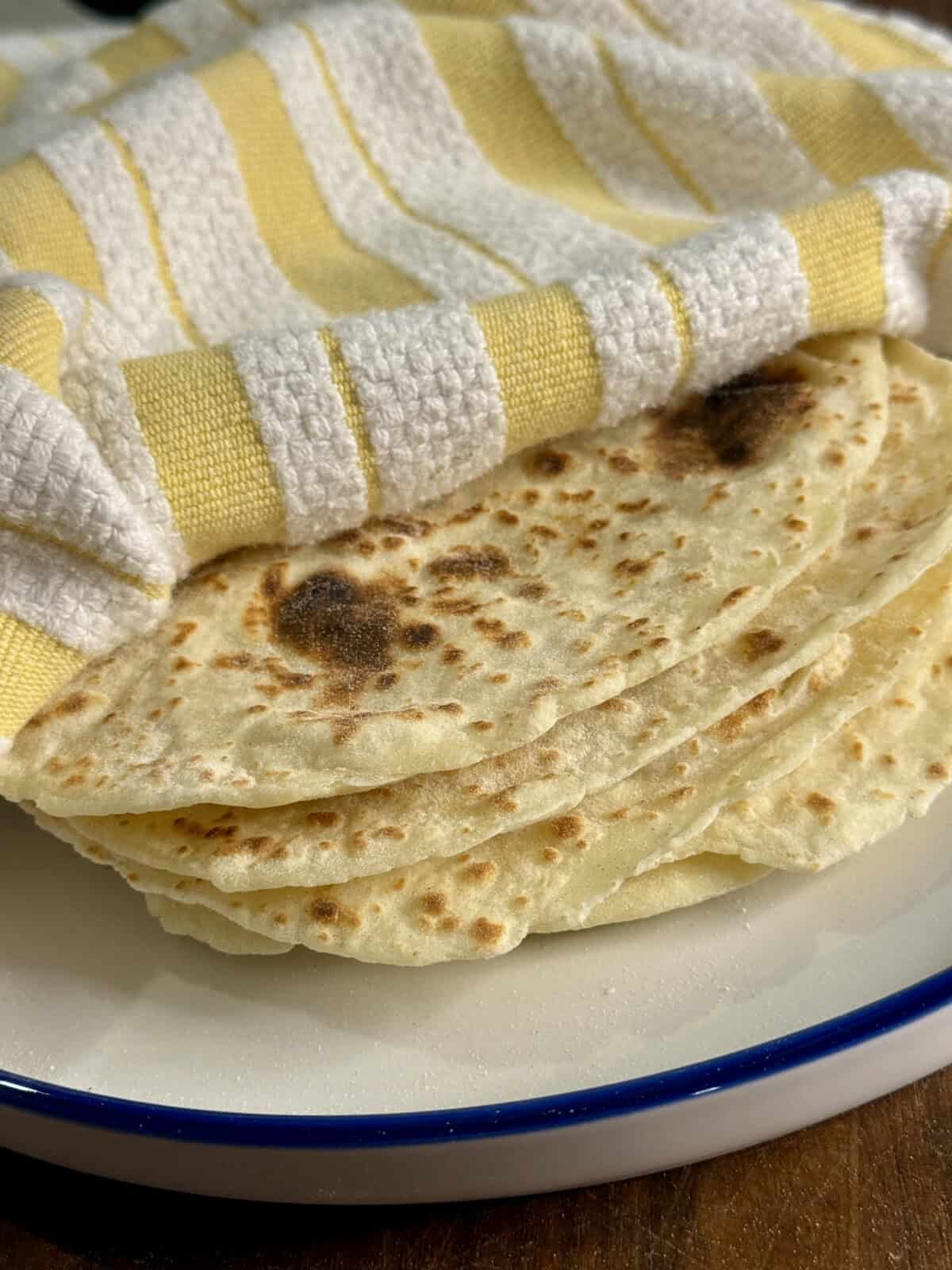 A stack of homemade tortillas sits on a white plate with a blue rim, partially covered by a yellow and white striped cloth.