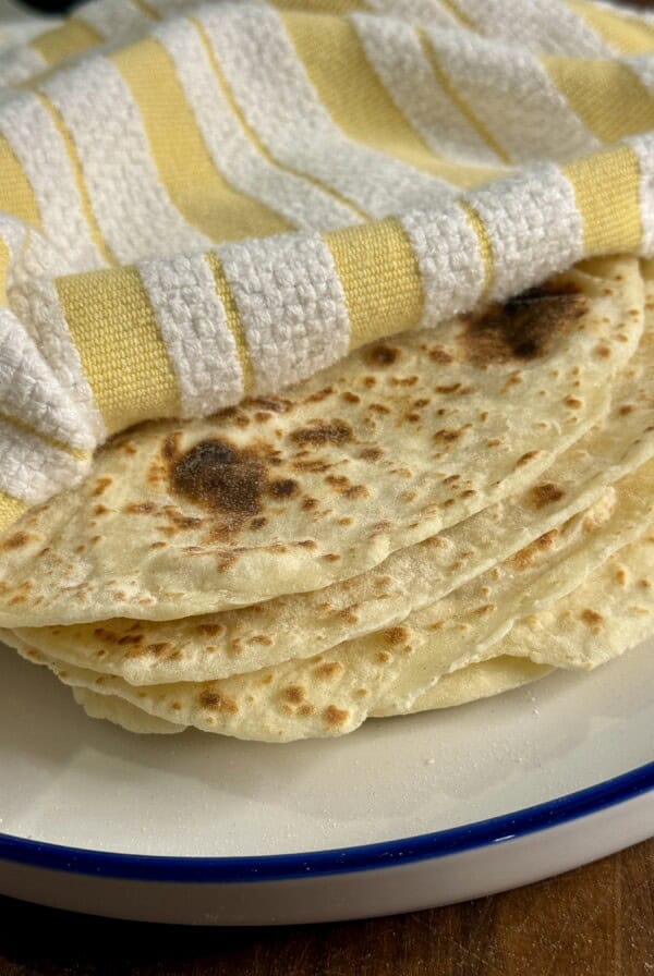 A stack of homemade tortillas sits on a white plate with a blue rim, partially covered by a yellow and white striped cloth.