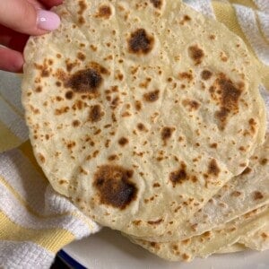 A hand holds up a freshly cooked, round flatbread with brown spots, above a plate stacked with more flatbreads. A yellow and white striped towel is in the background.