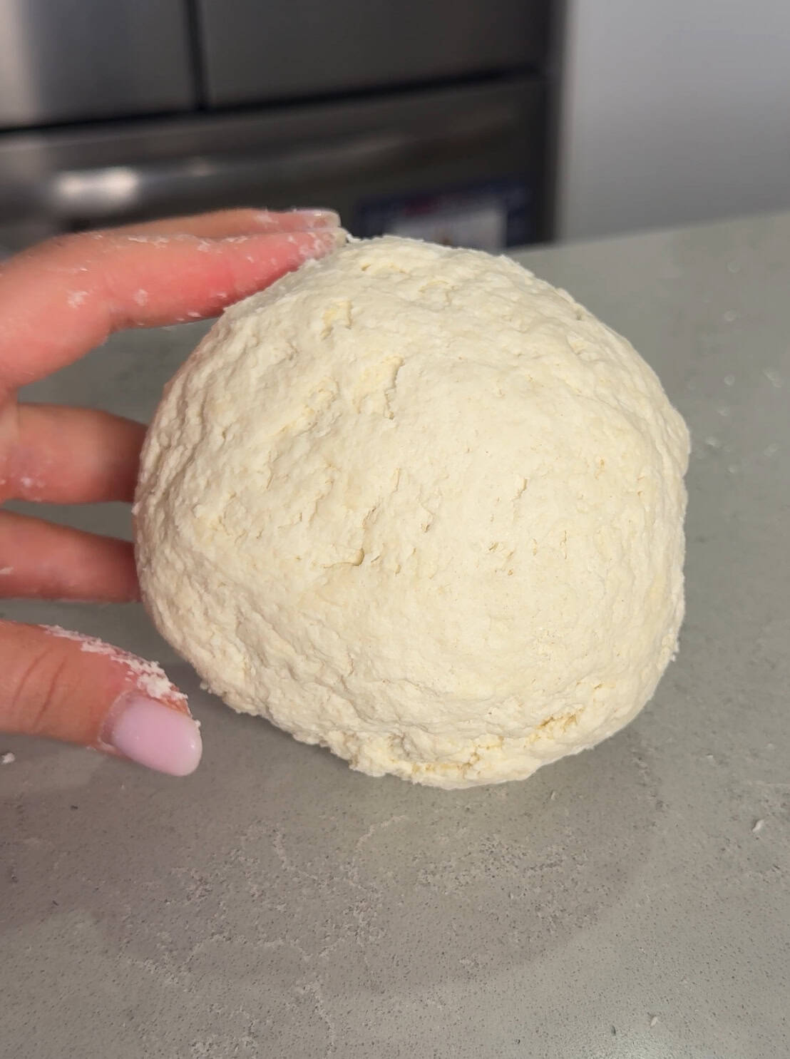 A hand with pink-painted nails is holding a rough, round ball of raw dough on a gray surface.