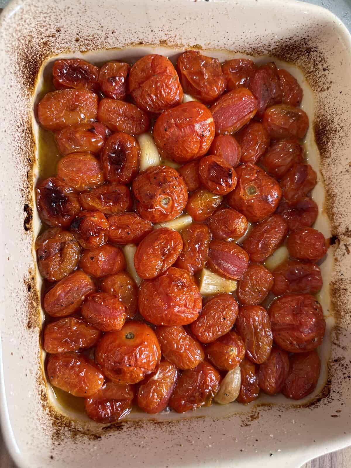 Roasted cherry tomatoes and garlic cloves in a white baking dish, with caramelized edges and a glossy, slightly wrinkled appearance from being cooked in olive oil.