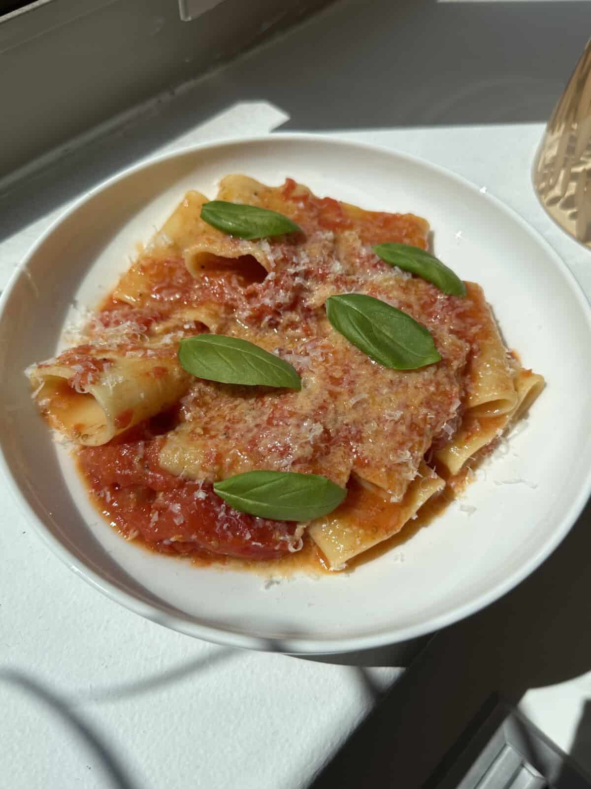 A white bowl of pasta with tomato sauce, topped with grated cheese and fresh basil leaves, sits on a white surface in bright sunlight.