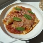 A white bowl of pasta with tomato sauce, topped with grated cheese and fresh basil leaves, sits on a white surface in bright sunlight.