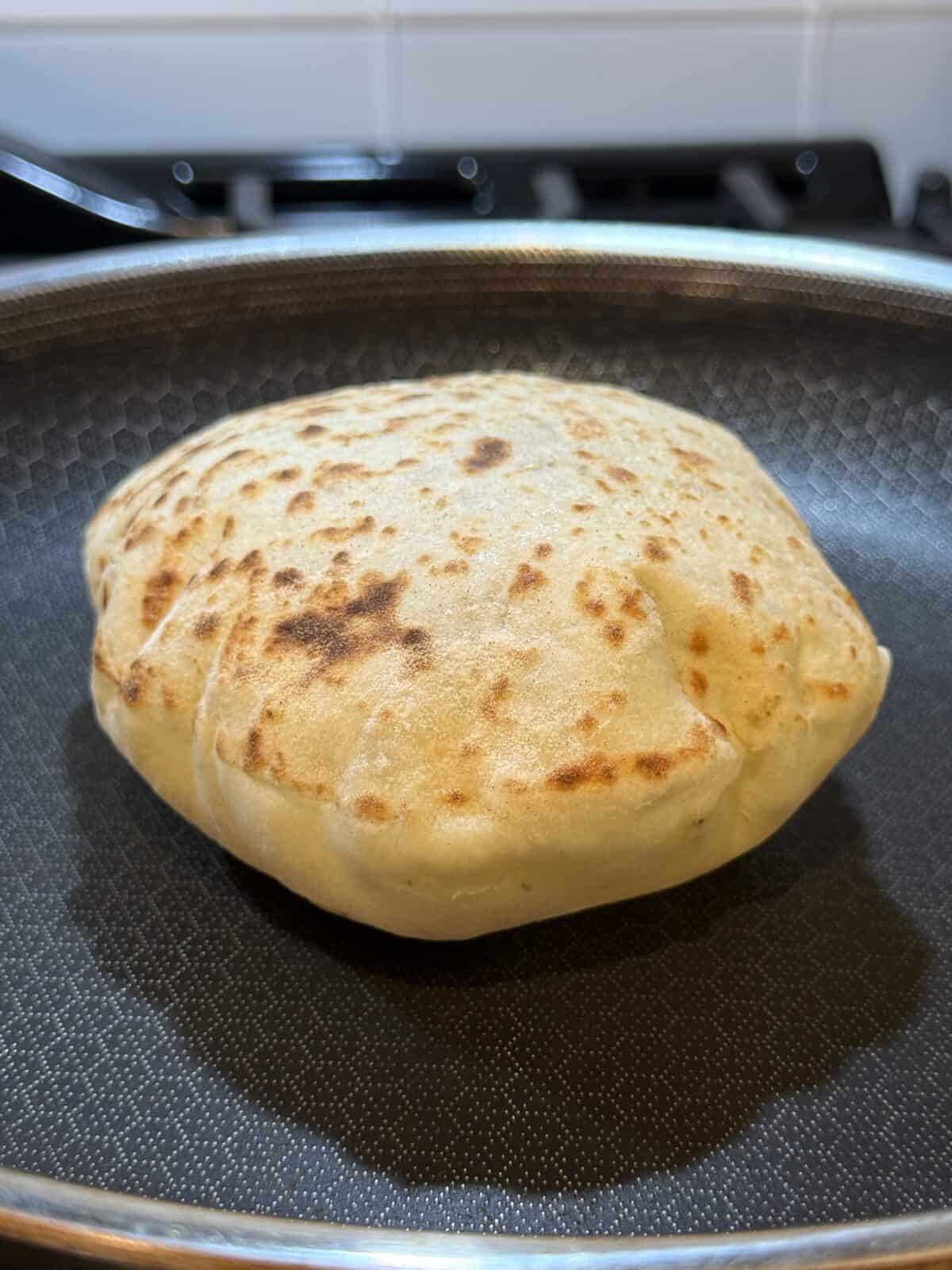 A golden-brown stuffed flatbread is cooking in a nonstick frying pan on a stovetop, with visible grill marks and a puffy texture.