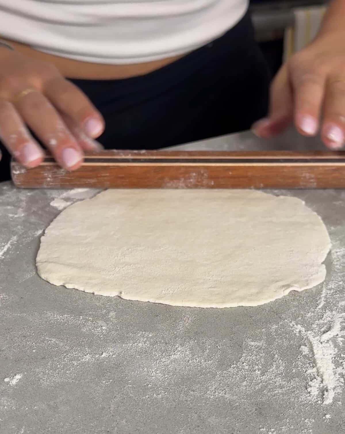 Hands rolling out dough with a wooden rolling pin on a floured surface, preparing it for baking.