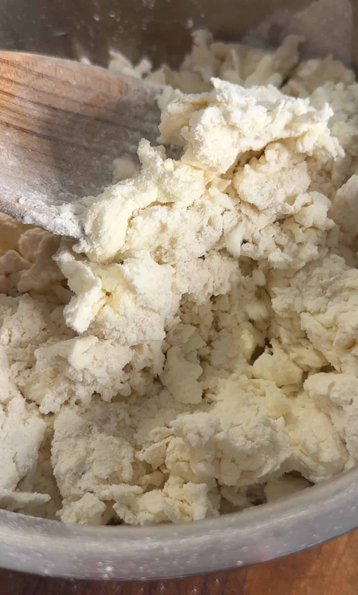 A close-up of rough, crumbly dough being mixed with a wooden spoon, showing uneven texture and pale yellow color.