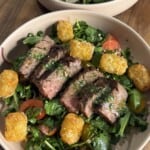 A bowl filled with sliced steak on a bed of mixed greens, cherry tomatoes, red onions, and topped with golden tater tots, all set on a wooden table.