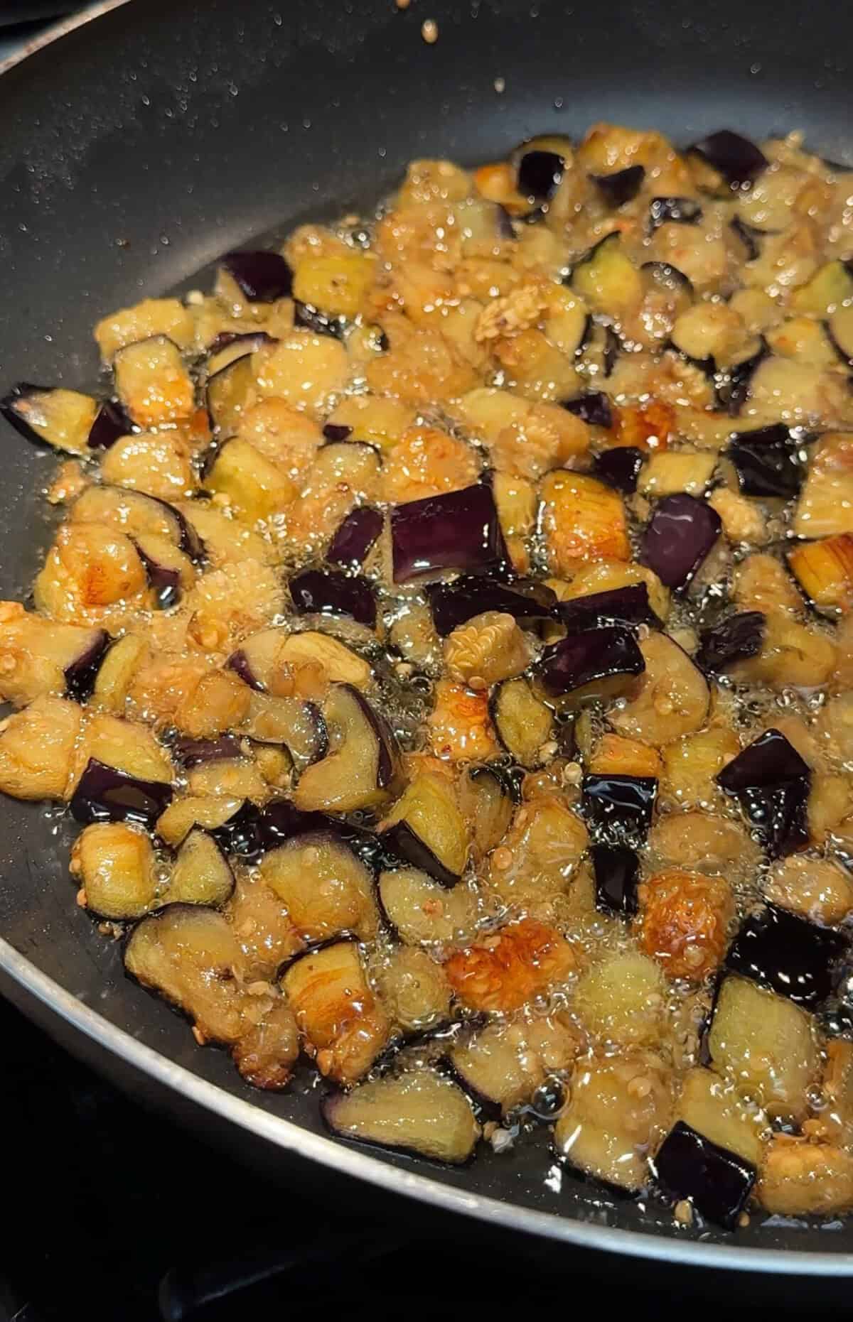 Diced pieces of eggplant frying in oil in a pan, with golden brown and purple pieces visible, and bubbles forming around the vegetables.