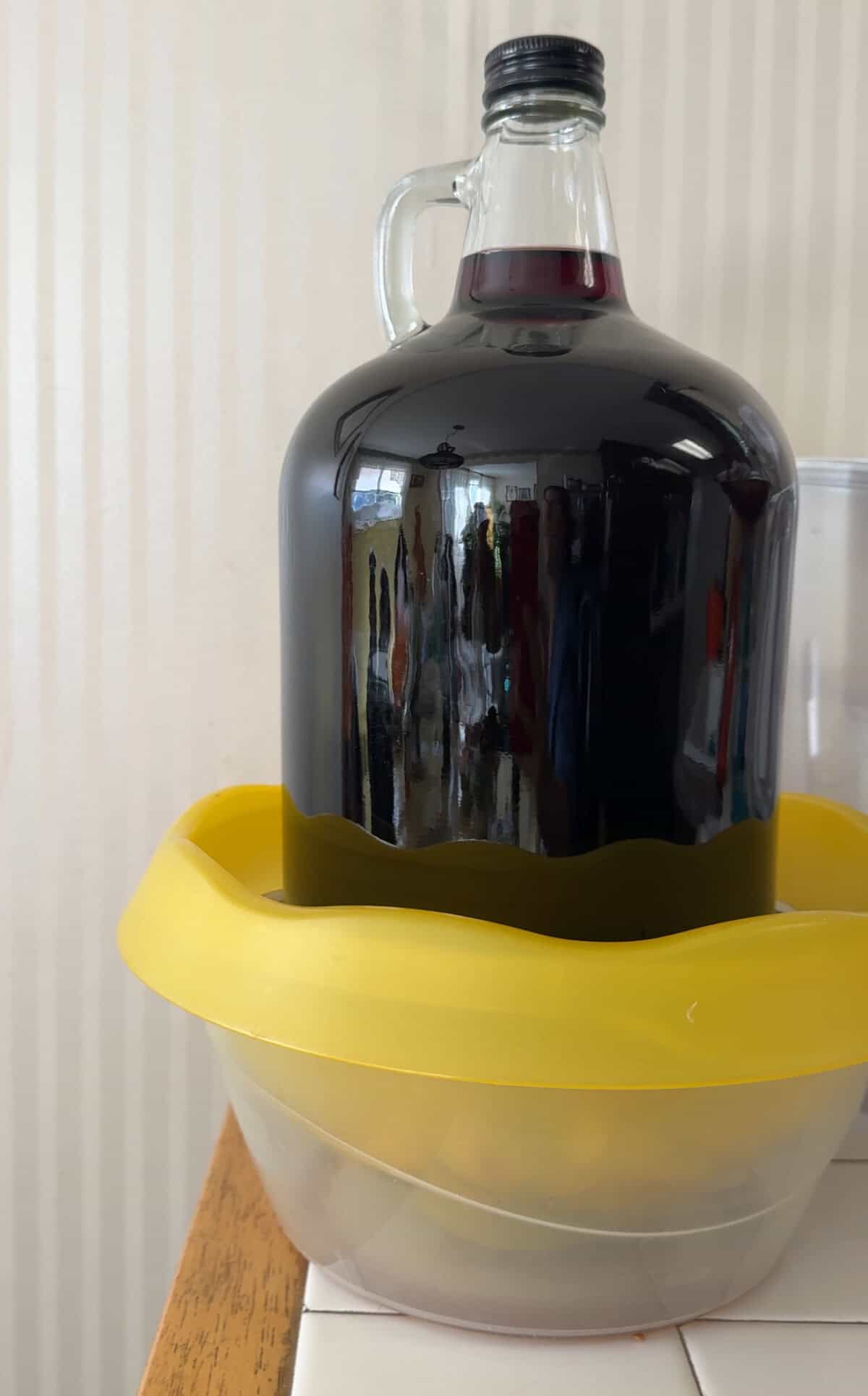 A large dark glass jug sits upside down in a yellow plastic ring, balanced on a clear bowl. The background features beige vertical stripes.