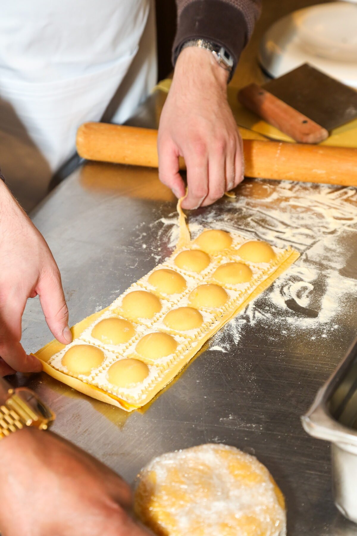 A person makes ravioli by pressing a sheet of pasta over round fillings on a floured metal countertop, using a rolling pin and a ravioli mold. Another hand holds dough nearby.
