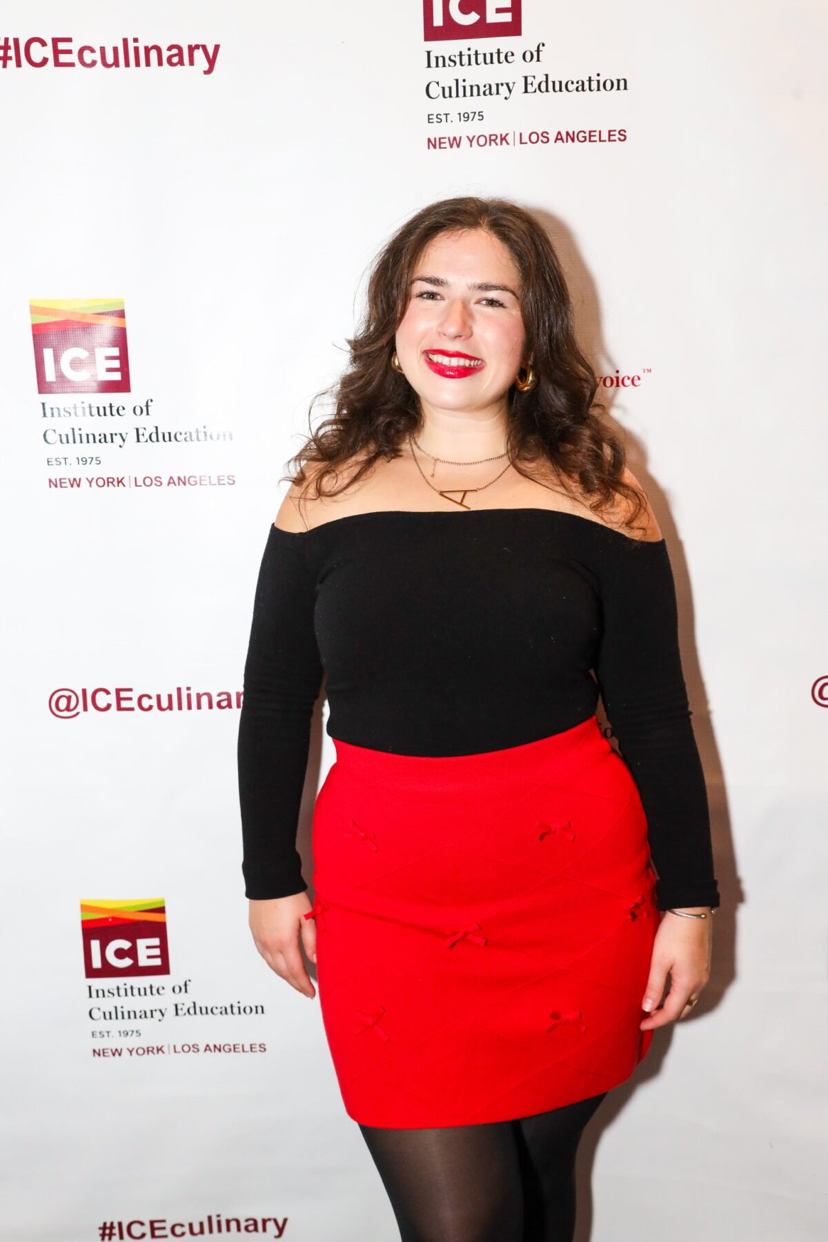 A woman with long brown hair smiles, wearing a black off-the-shoulder top and a red skirt. She stands in front of a step-and-repeat banner for the Institute of Culinary Education.