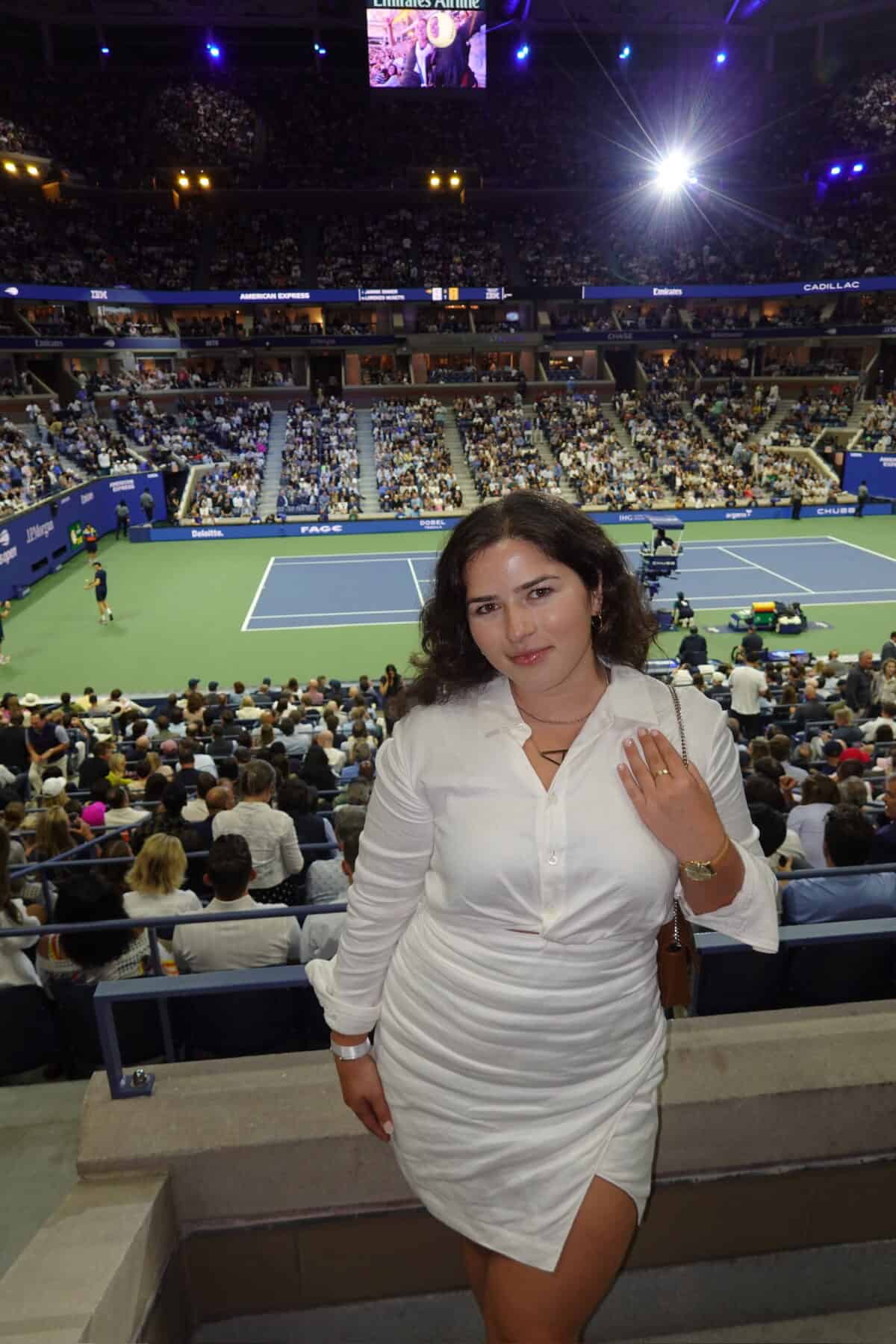 A woman in a white dress poses and smiles at a tennis stadium, with a large crowd and tennis match happening on the court behind her under bright lights.