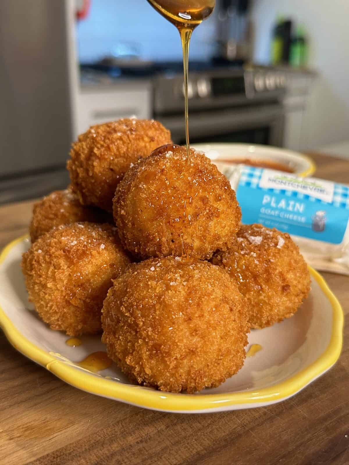 A plate of golden, crispy fried balls being drizzled with honey, with a package labeled plain goat cheese and a kitchen in the background.