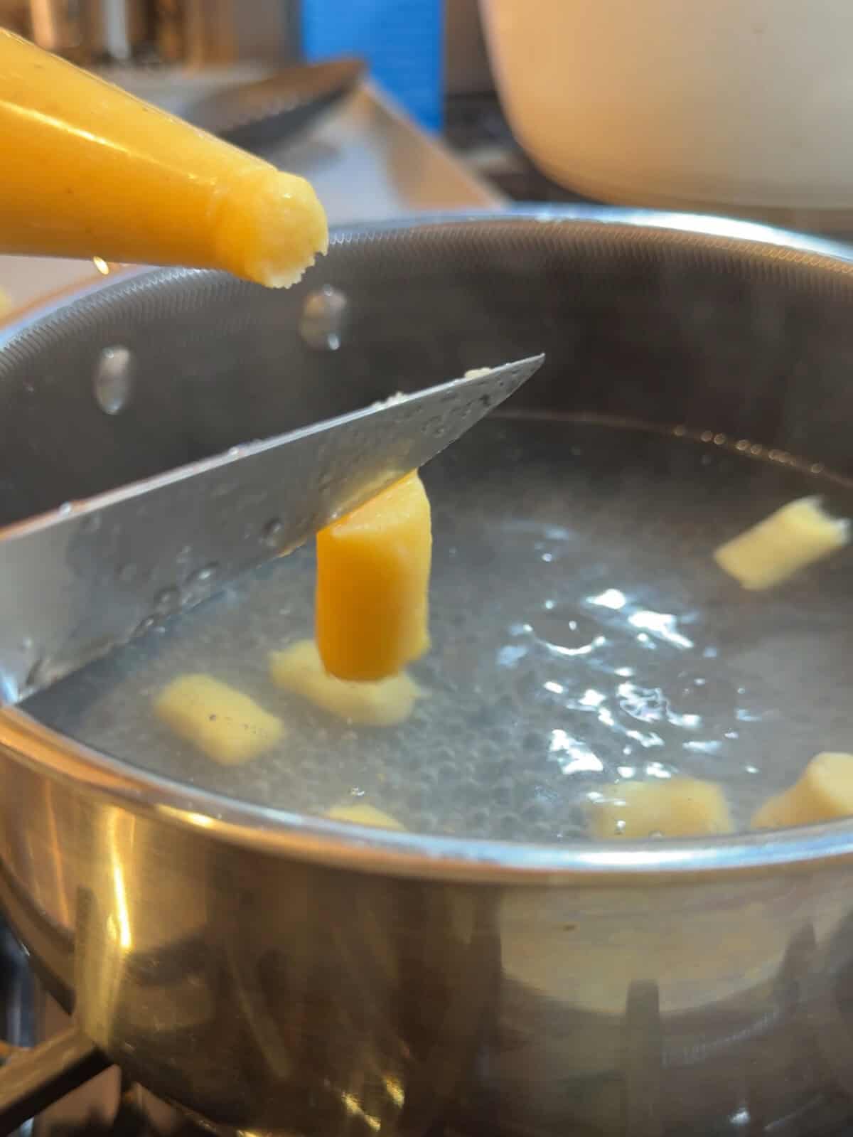 A knife slices yellow dough over a pot of boiling water, with pieces of dough floating inside the pot.