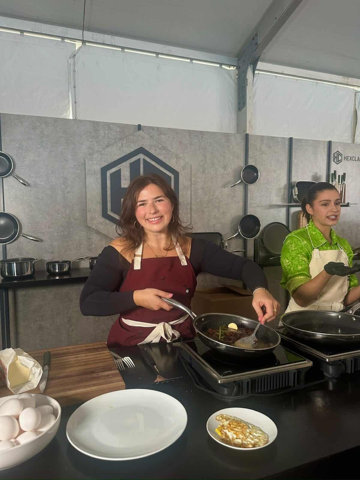 Two women cooking at a kitchen demo. The woman in front, wearing a red apron, is smiling while cooking food in a frying pan, while the woman in green behind her prepares food. Eggs and plates are on the counter.
