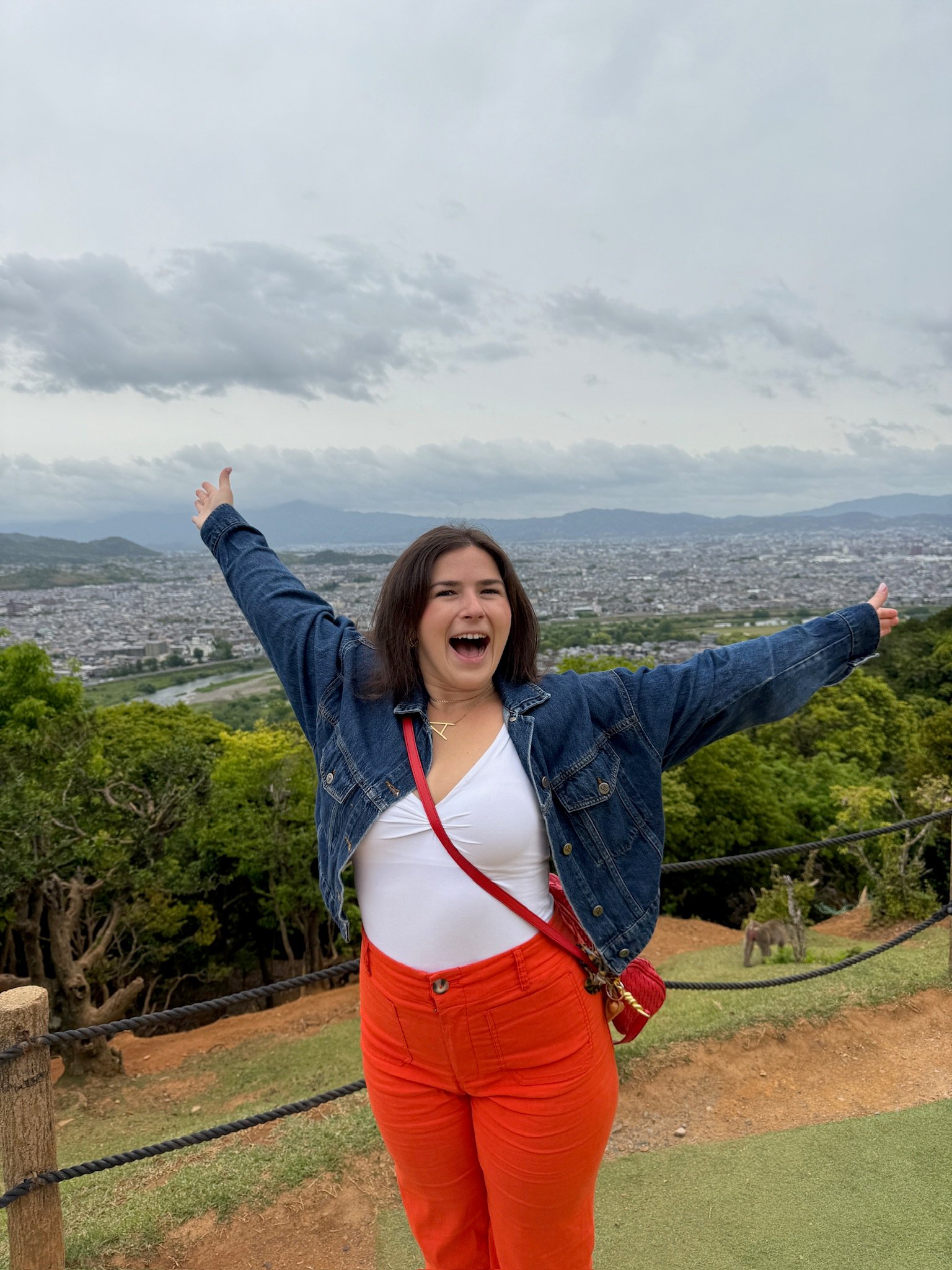 A woman in a white top, red pants, and denim jacket stands outdoors with arms outstretched, smiling. Behind her is a scenic view of a sprawling city, green trees, and cloudy sky.