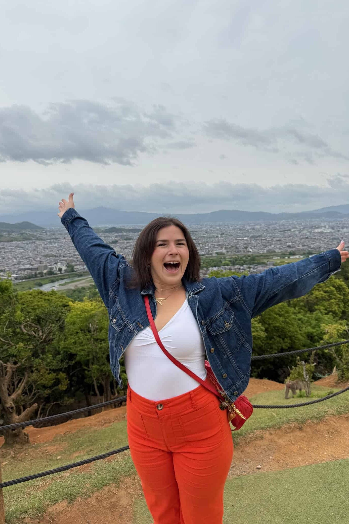 A woman with short brown hair, wearing a denim jacket, white top, and bright orange pants, stands outdoors with arms outstretched, smiling, with a city and mountains visible in the background.