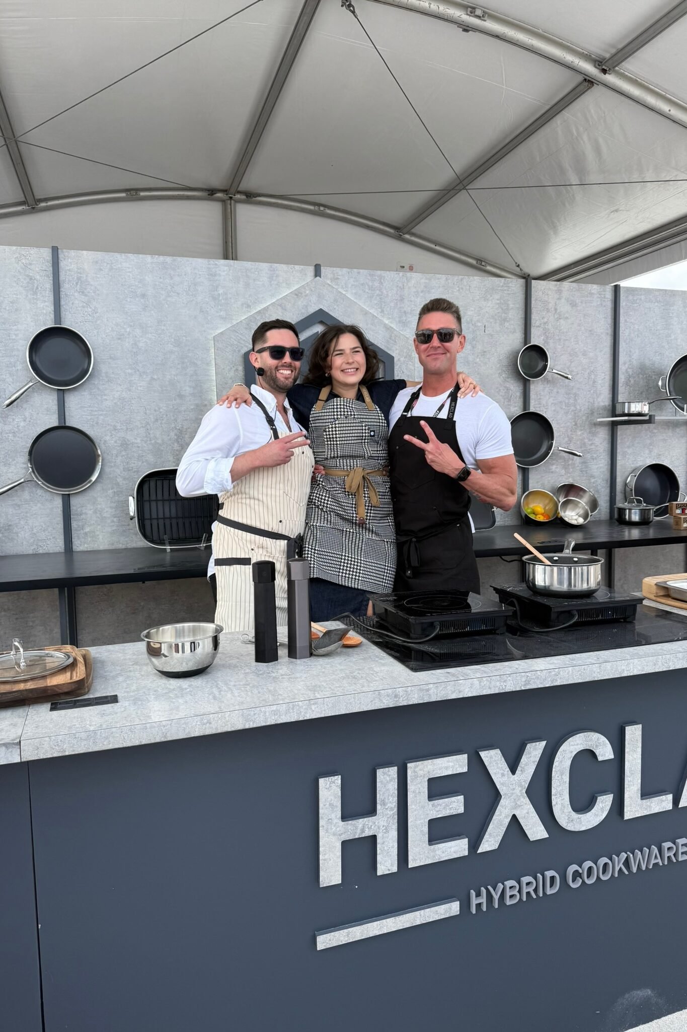 Three people wearing aprons stand smiling in front of a demonstration kitchen with pans on the wall behind them. They are posing under a tent at a HEXCLAD Hybrid Cookware event. One person makes a peace sign.