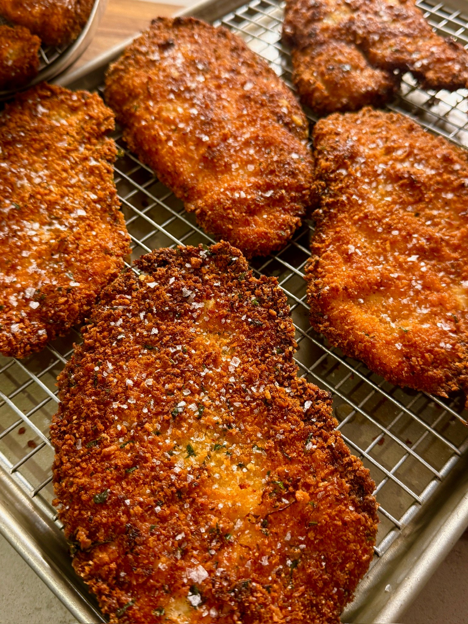 Golden, crispy breaded cutlets topped with coarse salt rest on a wire rack over a baking sheet, fresh from frying and ready to serve.