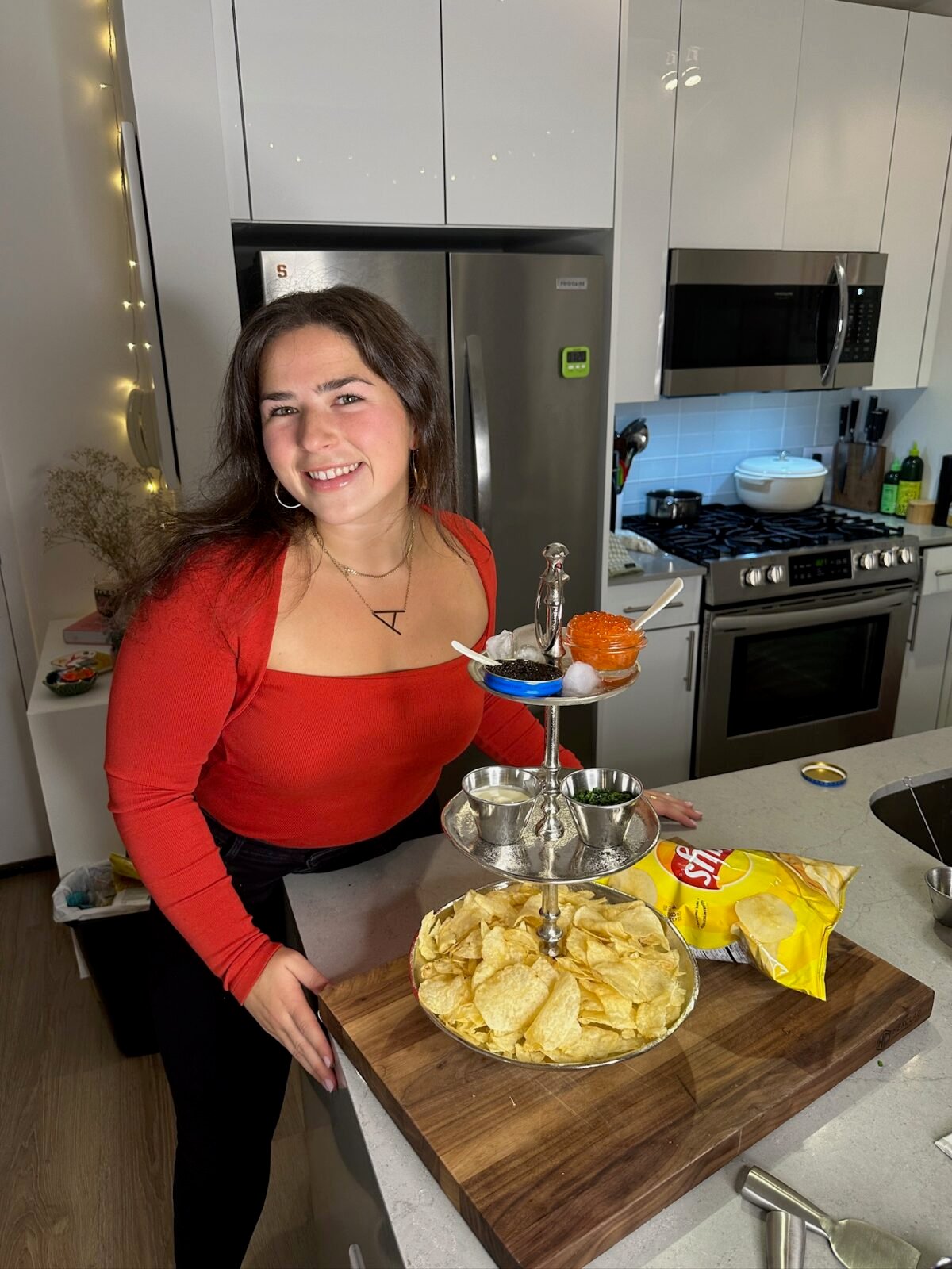 A woman in a red top smiles while standing in a modern kitchen beside a tiered tray with potato chips, blue dip, and red caviar. A bag of Lays chips and kitchen appliances are visible in the background.
