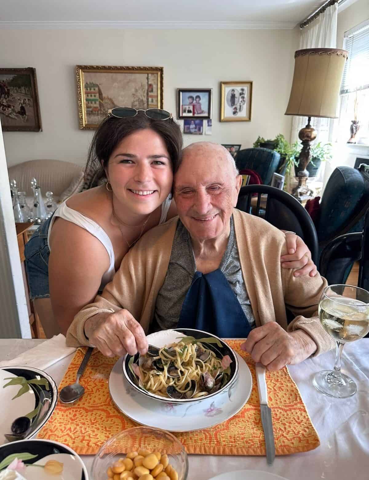 A young woman stands, smiling and hugging an elderly man who is seated at a dining table with a bowl of pasta. Both are smiling at the camera. The table is set with dishes and a glass of white wine. Artwork decorates the wall behind them.