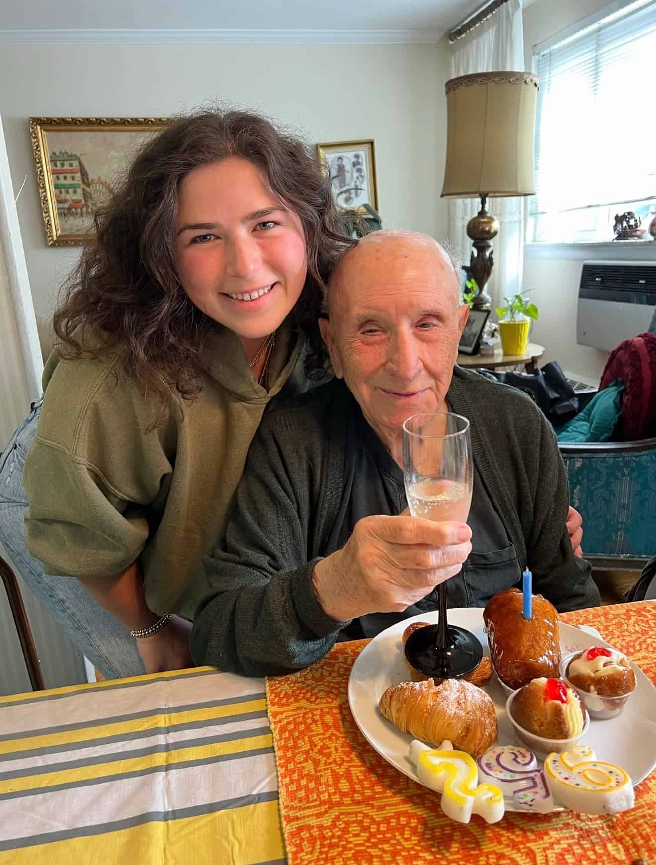A smiling young woman stands next to an elderly man holding a glass and sitting at a table with pastries, cakes, and a birthday candle. They appear happy, celebrating in a cozy, well-lit room.