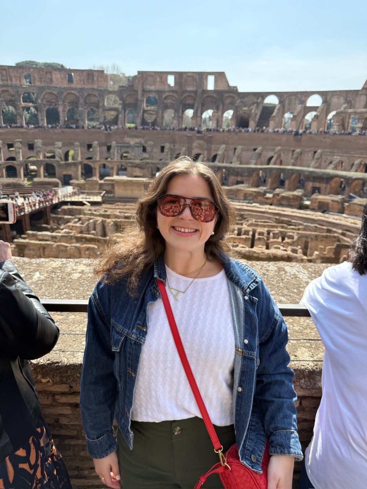A woman wearing sunglasses, a denim jacket, and a red purse smiles while standing in front of the ancient Roman Colosseum on a sunny day.
