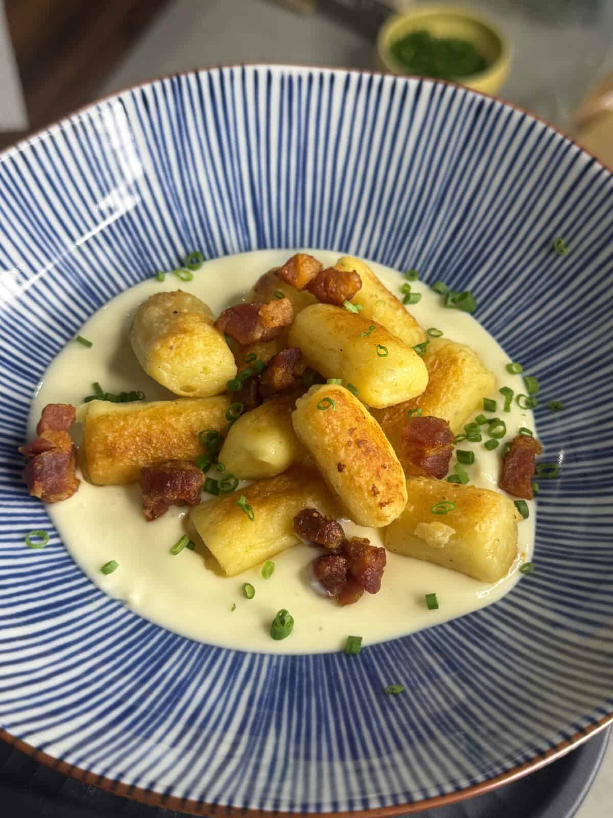 Golden-brown gnocchi on a creamy white sauce, topped with crispy bacon pieces and chopped chives, served in a blue and white striped bowl.