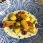 Golden-brown gnocchi on a creamy white sauce, topped with crispy bacon pieces and chopped chives, served in a blue and white striped bowl.