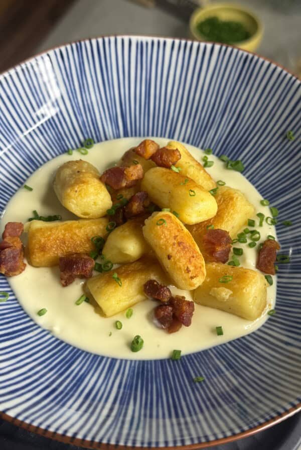 Golden-brown gnocchi on a creamy white sauce, topped with crispy bacon pieces and chopped chives, served in a blue and white striped bowl.