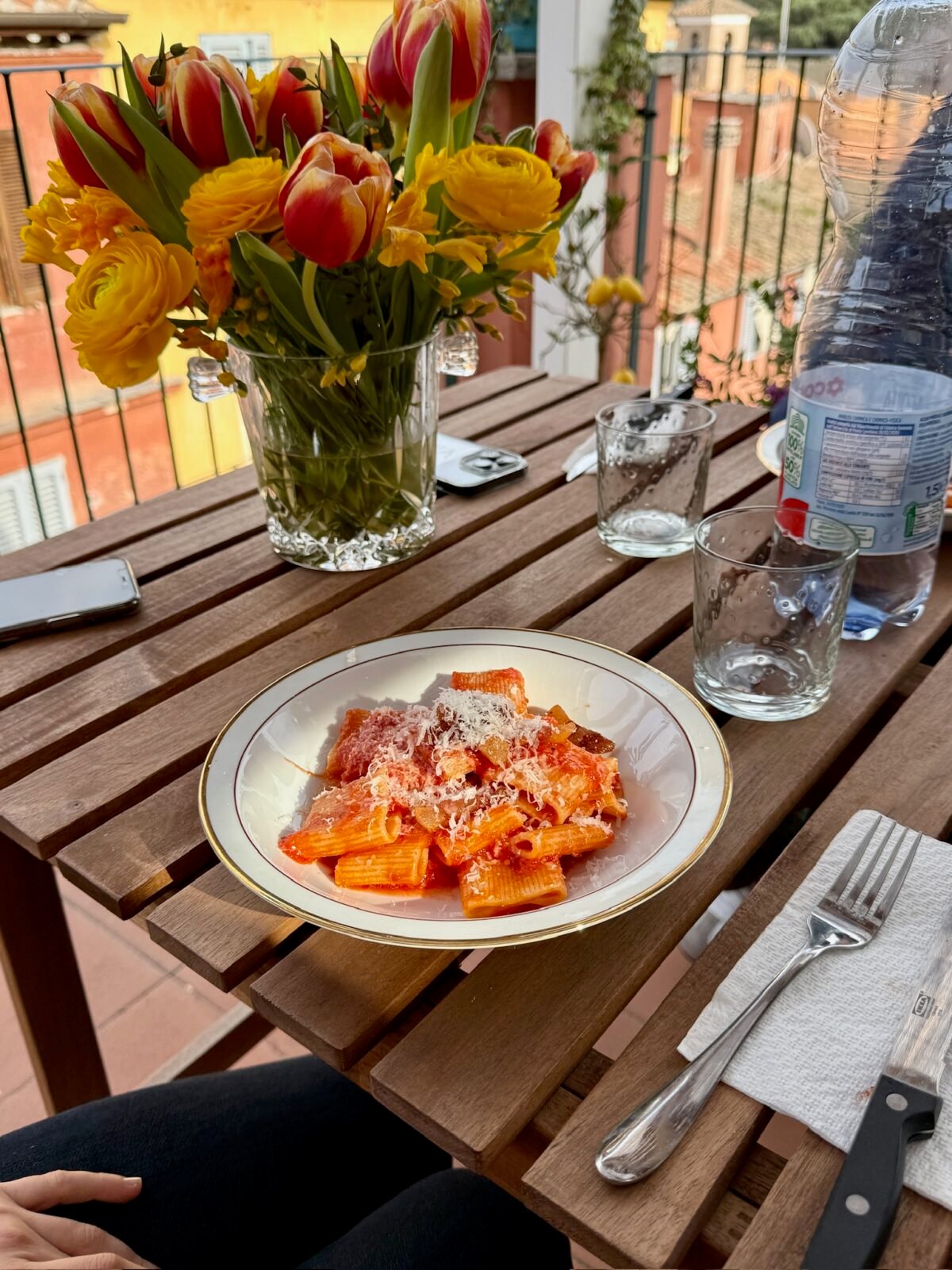 A wooden outdoor table set with a plate of pasta in tomato sauce, two empty glasses, a large bottle of water, a fork on a napkin, and a vase of yellow and red flowers. Rooftops are visible in the background.
