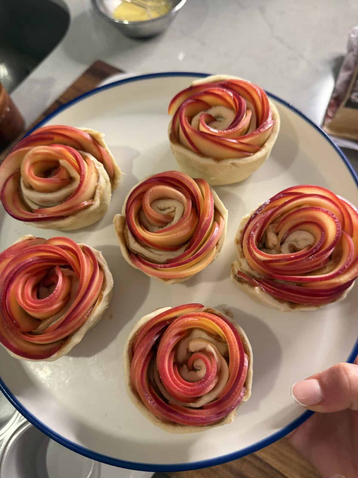A hand holds a white plate with six pastries shaped like roses, made from thinly sliced red and pink apples arranged in spirals atop a light dough base. A kitchen counter with ingredients is visible in the background.
