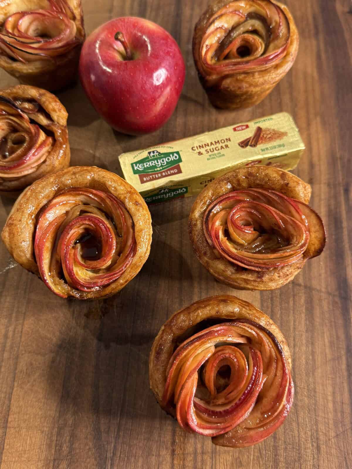 Apple rose pastries are arranged on a wooden surface with a red apple and a pack of Kerrygold Cinnamon & Sugar Butter in the background. The pastries have thinly sliced apples forming a rose shape.