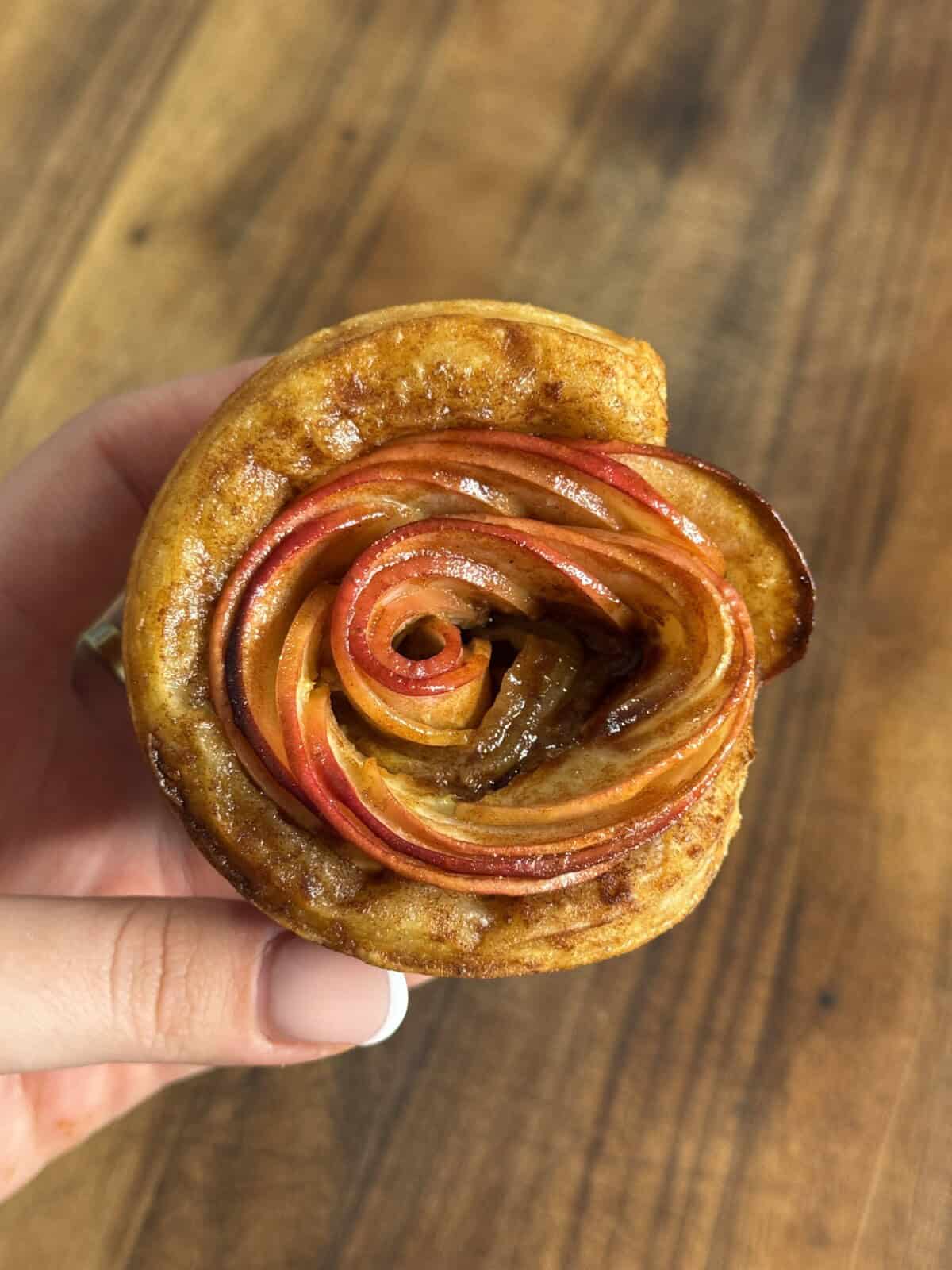 A hand holds a pastry shaped like a rose, made from thinly sliced red apples arranged in spirals, with a golden-brown crust, against a wooden background.