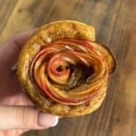 A hand holds a pastry shaped like a rose, made from thinly sliced red apples arranged in spirals, with a golden-brown crust, against a wooden background.
