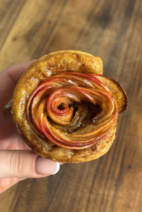 A hand holds a pastry shaped like a rose, made from thinly sliced red apples arranged in spirals, with a golden-brown crust, against a wooden background.