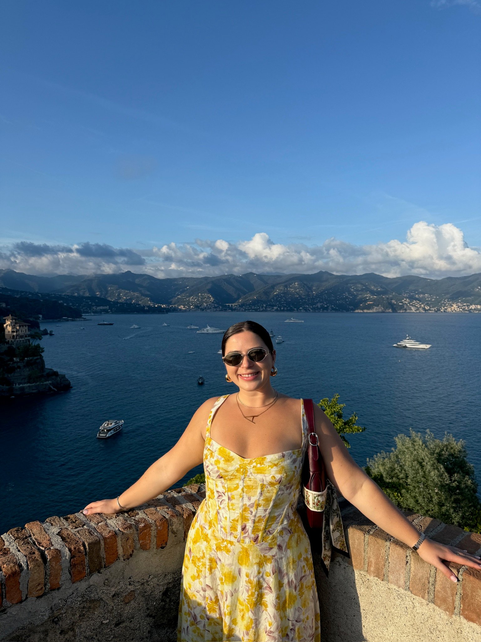 A woman in a yellow floral dress and sunglasses smiles while standing by a stone wall, overlooking a scenic bay with boats and distant mountains under a blue sky.