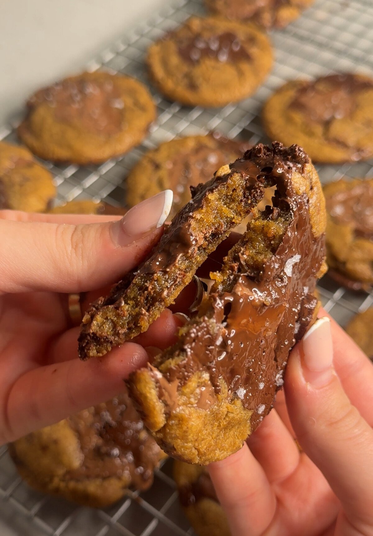 A person breaks apart a gooey chocolate chip cookie with melted chocolate in the center, while more cookies cool on a wire rack in the background.