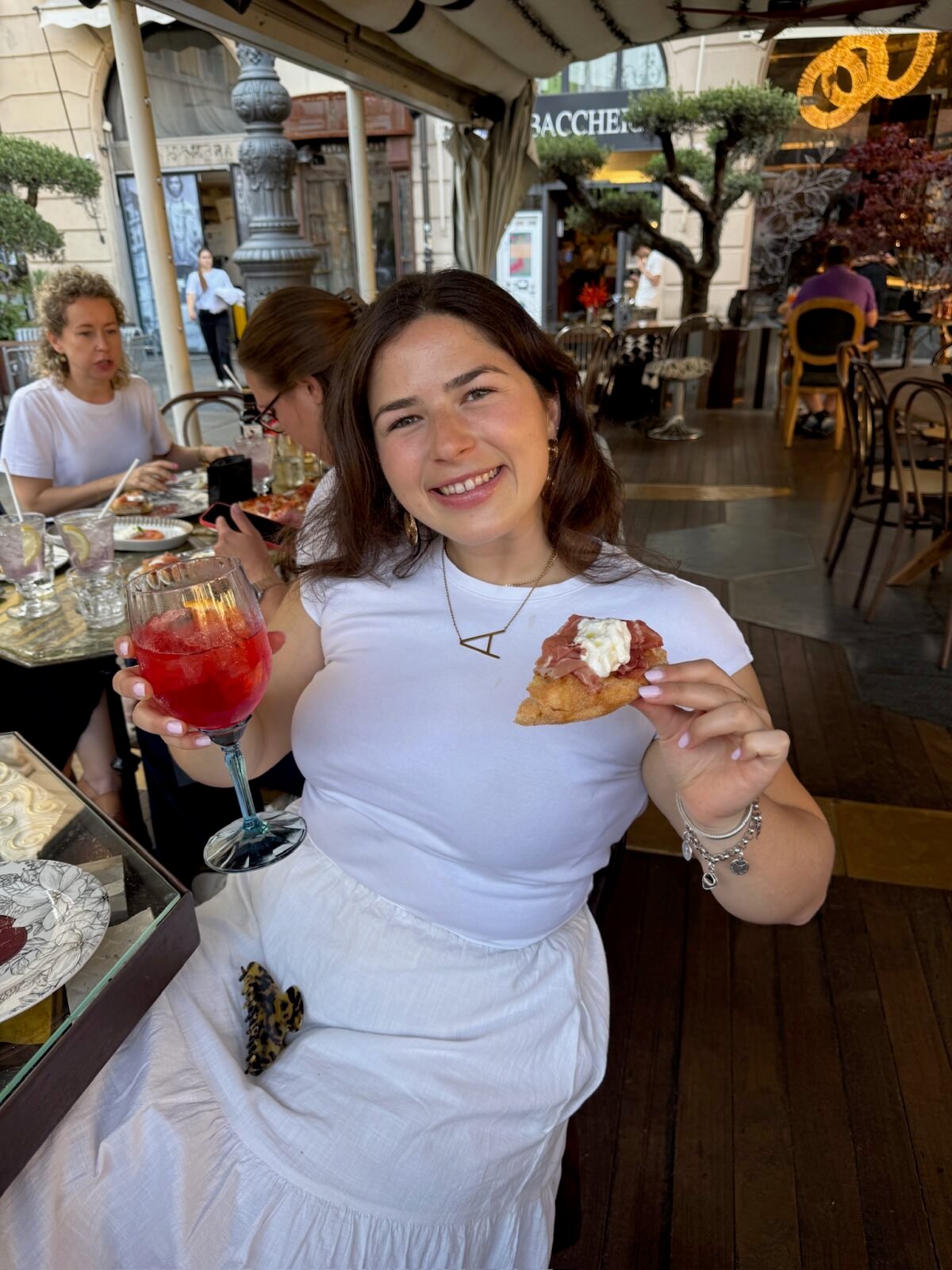 A smiling woman in a white shirt and skirt sits at a restaurant table, holding a red drink in one hand and a piece of bread with toppings in the other. Other diners and a stylish indoor-outdoor setting are visible behind her.